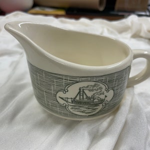 May include: Cream-colored gravy boat with a black and white illustration of a steamboat. The boat is in a decorative frame, and the body of the boat is decorated with a crosshatch pattern. The gravy boat has a handle and a spout.