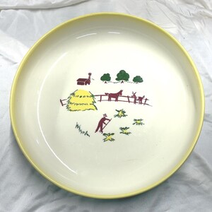 Peut inclure: Une assiette vintage de couleur crème avec un bord jaune. L'assiette présente une scène de ferme avec une grange, une botte de foin, une clôture et des personnages en rouge, vert et marron. L'assiette a un design classique et nostalgique.