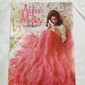 Peut inclure: Une couverture en crochet rose avec des franges, une femme est assise sur un lit avec la couverture drapée sur elle. La couverture du livre indique "Afghan Medley by Bernat" et "Book No. 168 Price $1.50".