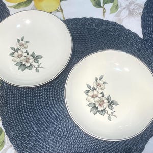 May include: Two white ceramic bowls with a floral design of white and pink flowers and grey leaves. The bowls have a dark grey trim and are on a blue woven placemat. The background has a floral pattern.