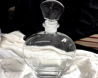 May include: A clear glass perfume bottle with a faceted, hexagonal base and a rounded body. The bottle has a clear, cylindrical neck and a clear stopper with a dark, faceted top. The bottle sits on a white cloth.