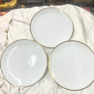 Op de afbeelding: Drie witte keramische borden met een gouden rand. De borden zijn rond en hebben een eenvoudig, elegant ontwerp. De borden zijn gerangschikt op een witte doek.