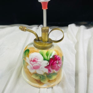 Könnte beinhalten: Eine kleine, porzellanene Gießkanne mit einem Messing-Ausguss und -Griff. Die Kanne ist mit einem Blumenmuster aus rosa und roten Rosen auf cremefarbenem Hintergrund verziert.