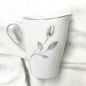 May include: White porcelain creamer with a silver rim and a handle. The creamer features a delicate illustration of a pink rose bud with gray leaves and stem. The creamer has a curved design and is suitable for serving cream or milk.