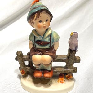 May include: A ceramic figurine of a young boy in traditional Bavarian attire, seated on a wooden fence. The boy wears a green hat, shirt, and brown lederhosen. A small bird perches on the fence. The figurine is on a round base.