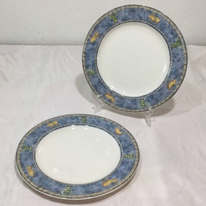 Puede incluir: Dos platos de cerámica blanca con un borde azul con un patrón de elementos decorativos amarillos y verdes. Los platos se exhiben sobre una superficie blanca, mostrando su diseño y combinación de colores. Los platos son redondos.