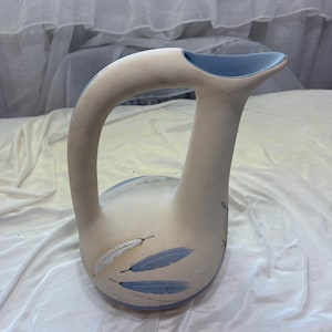 May include: A cream-colored ceramic pitcher with a curved handle and a blue interior. The pitcher is decorated with blue and white feather designs. The pitcher is likely used for serving beverages.