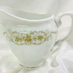 May include: White porcelain creamer with a floral design. The creamer features a spout, handle, and a gold trim. The floral pattern includes yellow roses and green leaves. A decorative piece for serving cream or other liquids.