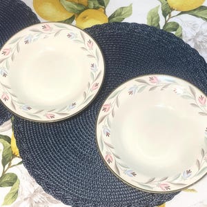 May include: Two cream-colored bowls with a floral design border featuring pink, blue, and gray flowers and green leaves. The bowls have a gold rim and are set on a dark blue placemat.