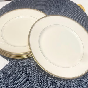 May include: A stack of off-white ceramic plates with a gold rim and a thin gold inner line. The plates are arranged on a dark blue placemat. The plates have a simple, elegant design.