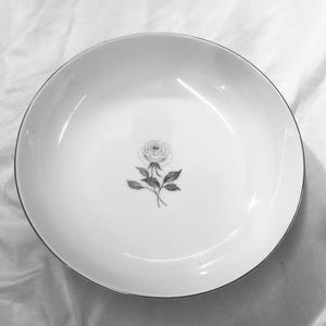 May include: A white ceramic serving bowl with a silver rim. The bowl features a delicate floral design of a rose with leaves in the center. The bowl is round and appears to be vintage.