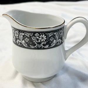 May include: White ceramic creamer with a black floral band and gold trim. The creamer has a handle and a spout for easy pouring. A decorative piece for serving cream or milk.