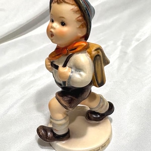 May include: A porcelain figurine of a young boy in a school uniform, with a backpack and a hat. The figurine is painted with a light skin tone, brown hair, and rosy cheeks. The boy is wearing a white shirt, brown shorts, and knee-high socks. The base reads "School Boy."
