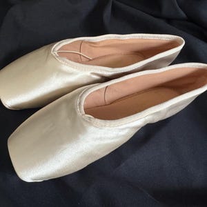 以下が含まれることがあります： ベージュのサテン素材のバレエシューズのペア。シューズは新品で、一度も履いていません。シューズは柔らかく快適な素材で作られており、クラシックなデザインです。