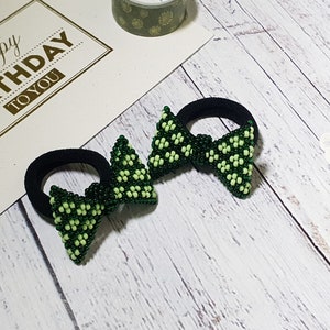 Puede incluir: Dos gomas para el pelo de cuentas verdes y blancas con bandas elásticas negras. Las cuentas están dispuestas en forma de lazo.