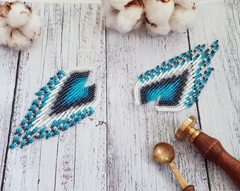 May include: A pair of long, beaded earrings with a geometric design in shades of white, blue, and gray. The earrings are hanging on a wooden surface.