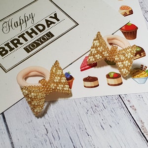 Puede incluir: Dos lazos para el pelo de cuentas doradas y blancas. Los lazos para el pelo están en una tarjeta blanca con el texto "Happy Birthday to You".