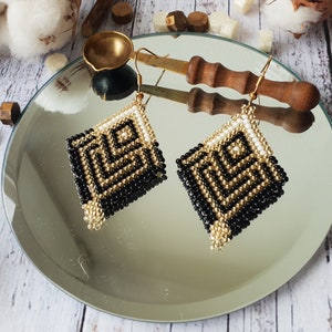 Puede incluir: Un par de pendientes geométricos de cuentas negras y doradas con un acento blanco. Los pendientes se muestran en una superficie de espejo.