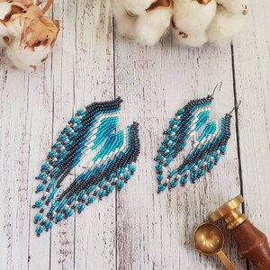 May include: A pair of long, beaded dangle earrings with a geometric pattern in shades of blue, black, white, and silver. The earrings are hanging from silver hooks.