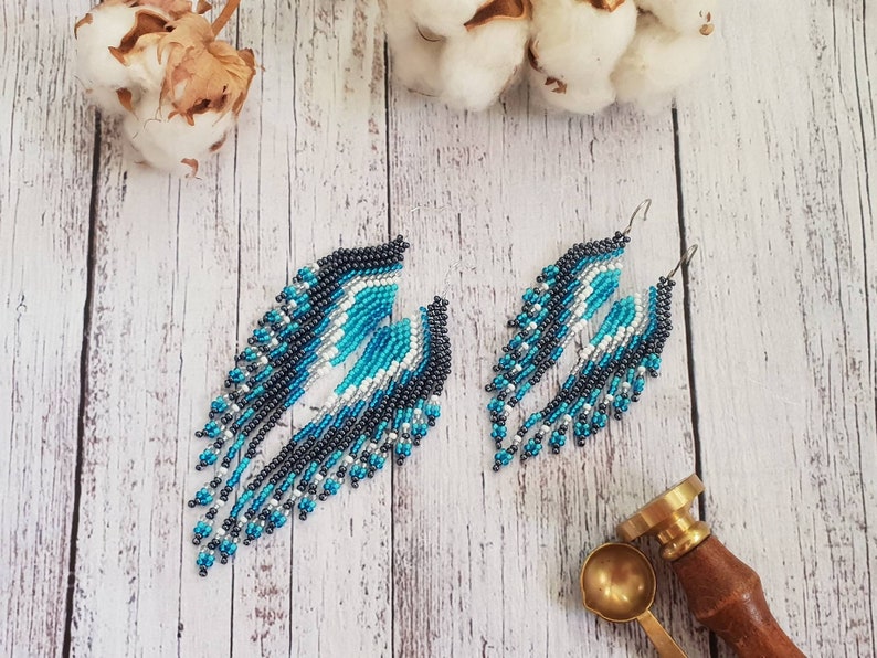 May include: A pair of long, beaded dangle earrings with a geometric pattern in shades of blue, black, white, and silver. The earrings are hanging from silver hooks.