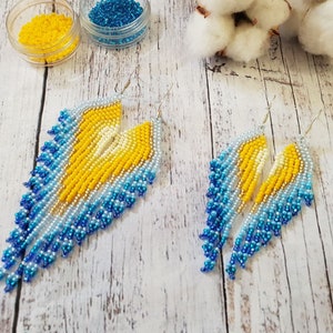 May include: A pair of long, beaded earrings with a gradient of blue, white and yellow beads. The earrings are teardrop shaped and dangle from silver hooks.