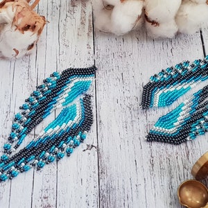 May include: A pair of long, beaded earrings with a geometric design in shades of blue, white, and gray. The earrings are made with small, seed beads and have a fringe-like effect.