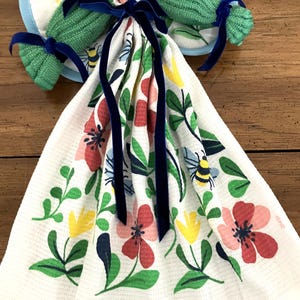 May include: A white kitchen towel with a floral pattern of red, pink, yellow, and blue flowers, green leaves, and bees. The towel is tied with a navy blue ribbon. Two green pot holders with blue trim and navy blue ribbons are also visible.