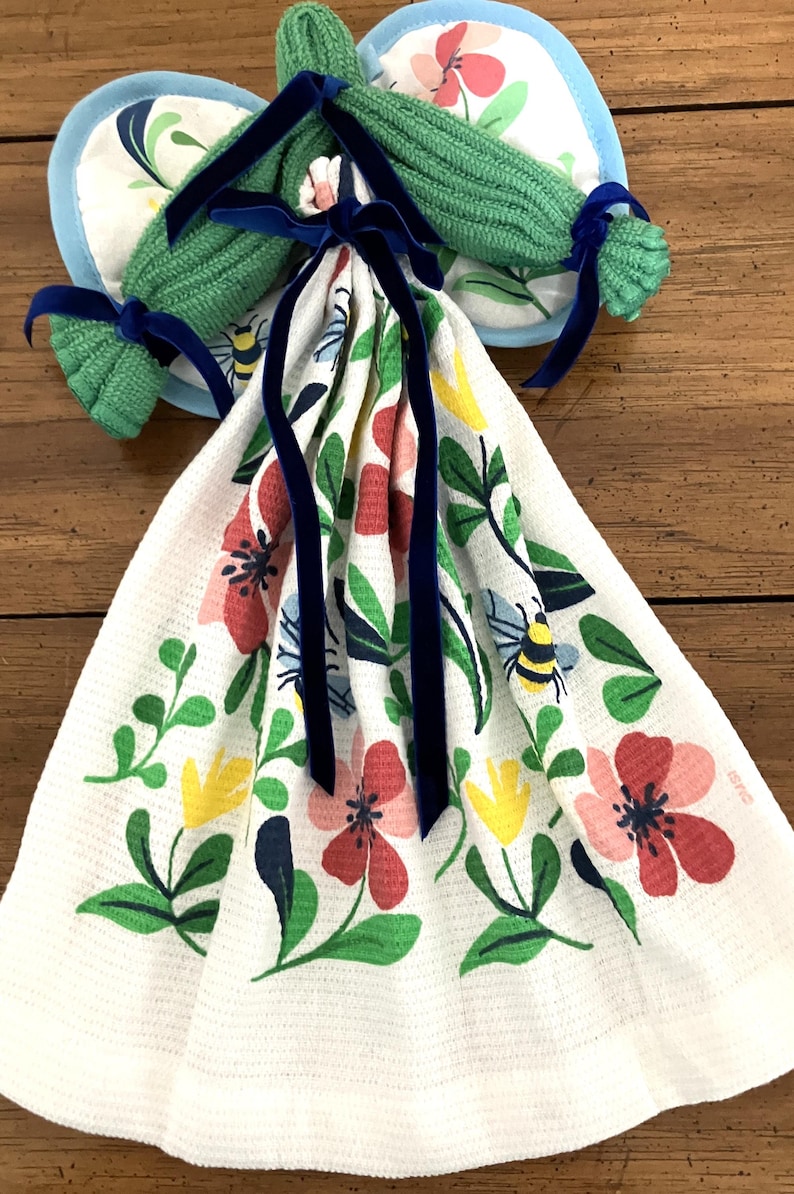 May include: A white kitchen towel with a floral pattern of red, pink, yellow, and blue flowers, green leaves, and bees. The towel is tied with a navy blue ribbon. Two green pot holders with blue trim and navy blue ribbons are also visible.