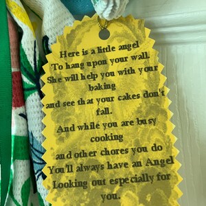 May include: A yellow, scalloped-edged tag with the text "Here is a little angel..." attached to a decorative kitchen towel. The towel has a floral pattern with green and red accents.