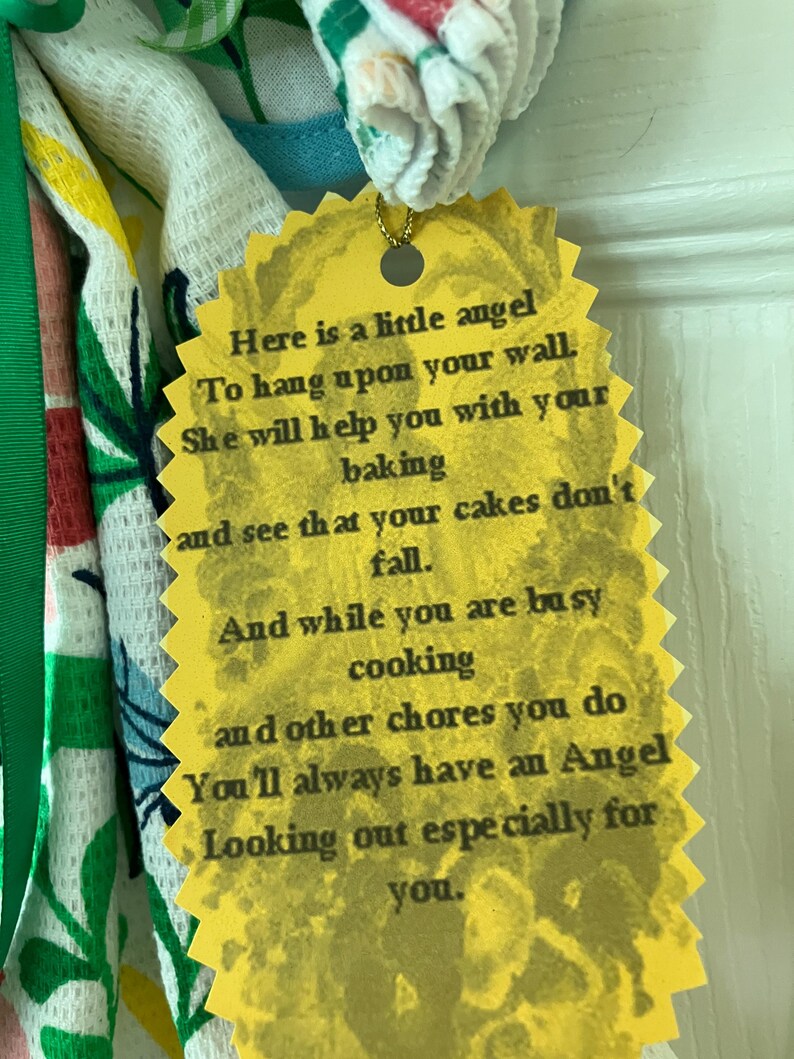 May include: A yellow, scalloped-edged tag with the text "Here is a little angel..." attached to a decorative kitchen towel. The towel has a floral pattern with green and red accents.