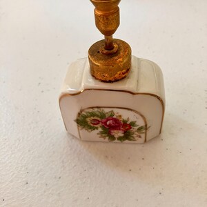 May include: A white porcelain perfume bottle with a rectangular shape and rounded corners. The front features a floral decal with red and pink roses and green leaves. It has a gold-toned metal pump mechanism with a weathered, aged finish.