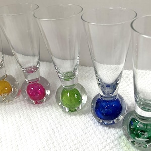May include: A set of five clear glass flutes, each featuring a distinctively shaped base containing a solid colored sphere. The spheres are yellow, pink, lime green, dark blue, and dark green, providing a vibrant contrast to the transparent glass.