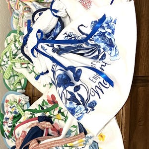 May include: A collection of folded white kitchen towels with various colorful floral and patriotic designs. Some towels feature blue floral patterns, while others display fireworks and stars. Each towel is tied with a coordinating ribbon.