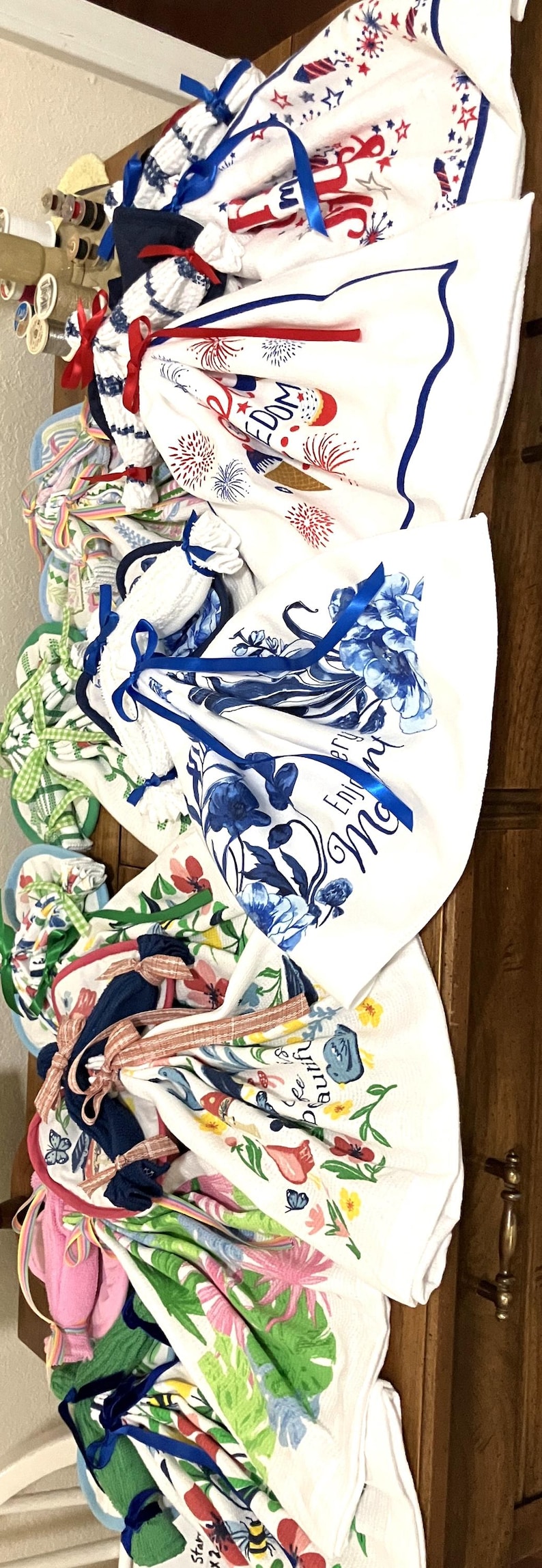May include: A collection of folded white kitchen towels with various colorful floral and patriotic designs. Some towels feature blue floral patterns, while others display fireworks and stars. Each towel is tied with a coordinating ribbon.