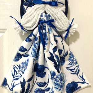 May include: A decorative angel-shaped kitchen towel. The angel features white terry cloth hair and wings, and a blue and white floral patterned towel for the dress. Blue satin ribbons accent the design. The wings are trimmed with a dark blue border.