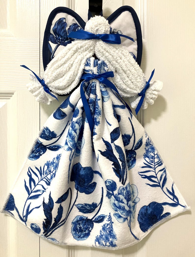 May include: A decorative angel-shaped kitchen towel. The angel features white terry cloth hair and wings, and a blue and white floral patterned towel for the dress. Blue satin ribbons accent the design. The wings are trimmed with a dark blue border.