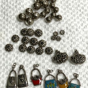 May include: A collection of metal jewelry components including smooth silver-toned spherical beads, antique silver-toned flower-shaped spacer beads, two ornate filigree charms, and six miniature handbag charms with colorful enamel accents.