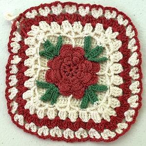 May include: A square crochet potholder featuring a central red rose with green leaves, surrounded by a cream-colored square pattern with a red border. The design includes a small loop at the top corner for hanging, crafted with yarn.
