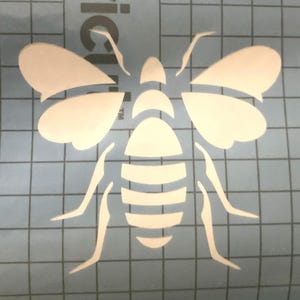 May include: White vinyl stencil of a bee with a striped body and wings. The stencil is on a blue grid background.