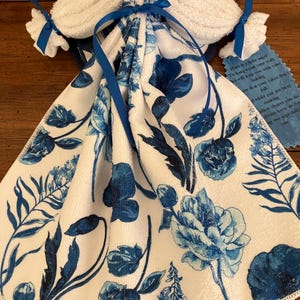 May include: A decorative towel set, featuring a white towel with a blue floral pattern. The set includes a folded towel with a bow and two smaller towels tied with blue ribbons. A small tag with text is attached.