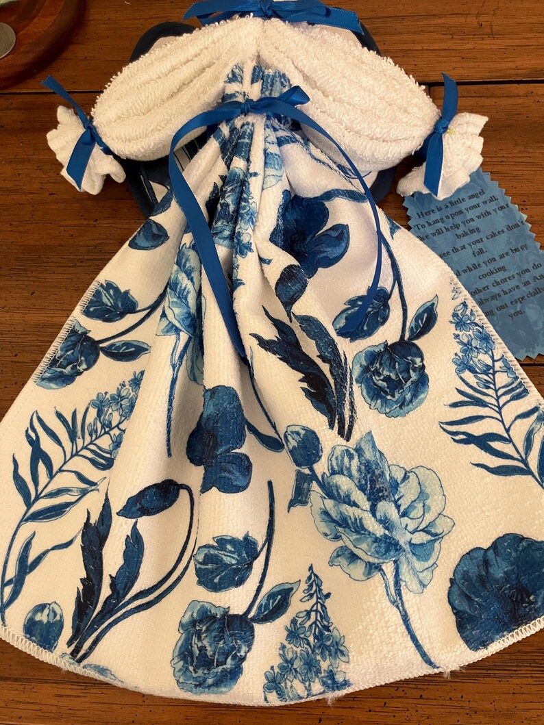 May include: A decorative towel set, featuring a white towel with a blue floral pattern. The set includes a folded towel with a bow and two smaller towels tied with blue ribbons. A small tag with text is attached.