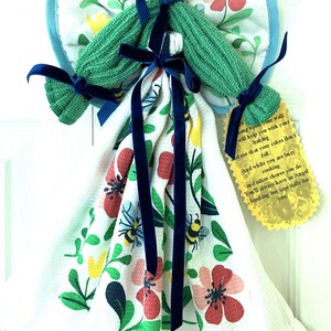 May include: An angel-shaped kitchen towel holder with a green knitted head and arms, blue ribbon accents, and a floral patterned towel. The wings are made of a blue-trimmed, floral-printed fabric. A yellow tag with text hangs from the towel.