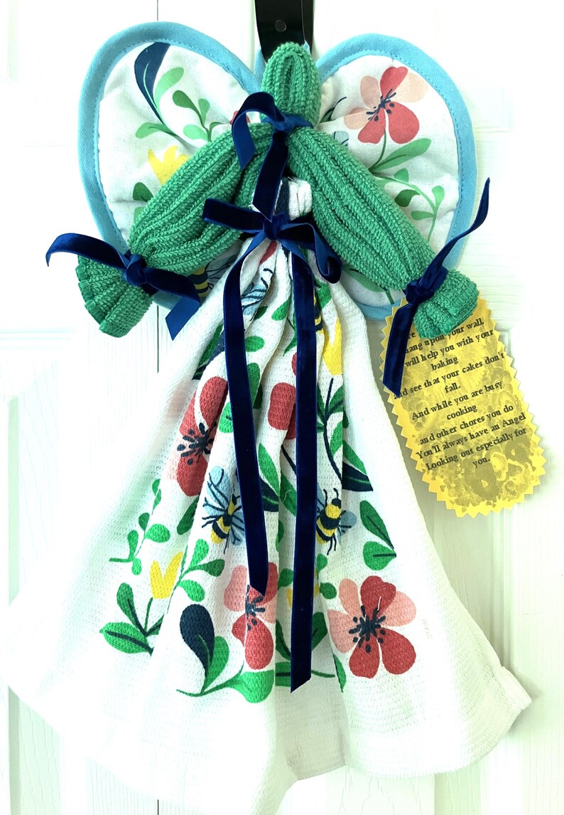 May include: An angel-shaped kitchen towel holder with a green knitted head and arms, blue ribbon accents, and a floral patterned towel. The wings are made of a blue-trimmed, floral-printed fabric. A yellow tag with text hangs from the towel.
