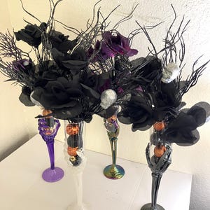 May include: Four tall champagne flutes with stems shaped like skeletal hands in purple, white, iridescent, and black. Each glass contains small orange pumpkins, black spheres, black roses, silver skulls, and glittery black twigs.