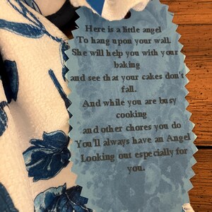 May include: A white and blue floral patterned kitchen towel with a blue tag. The tag has a scalloped edge and contains a poem about an angel helping with baking and chores.