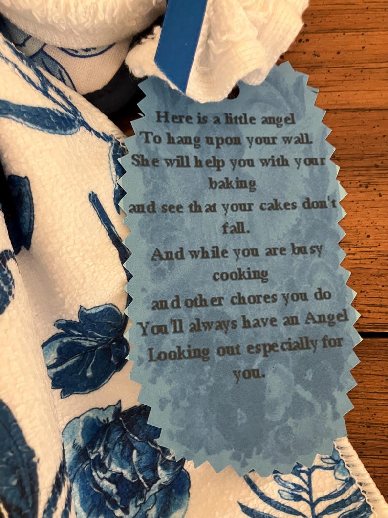 May include: A white and blue floral patterned kitchen towel with a blue tag. The tag has a scalloped edge and contains a poem about an angel helping with baking and chores.