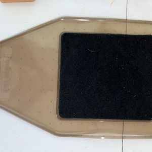 May include: A translucent brown plastic tray with a tapered, rounded shape and a small circular hole on the left side. A square, black, textured fabric pad is centered on the surface of the tray. The tray features a subtle grid of tiny dots.