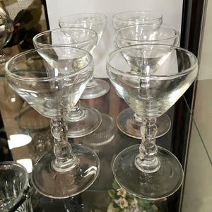 May include: A set of six clear glass stemmed goblets, each featuring a wide, rounded bowl and a stem decorated with three stacked spherical glass beads, resting on a circular base. The glassware is arranged in a group on a reflective surface.