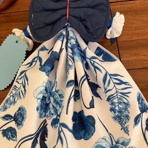 May include: A decorative kitchen towel with a blue floral pattern and a navy blue quilted bow-shaped top. The towel has a white background with blue flowers and leaves. The bow is attached with a red string.