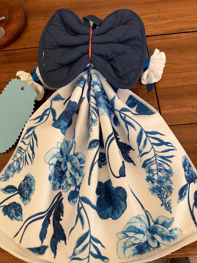 May include: A decorative kitchen towel with a blue floral pattern and a navy blue quilted bow-shaped top. The towel has a white background with blue flowers and leaves. The bow is attached with a red string.